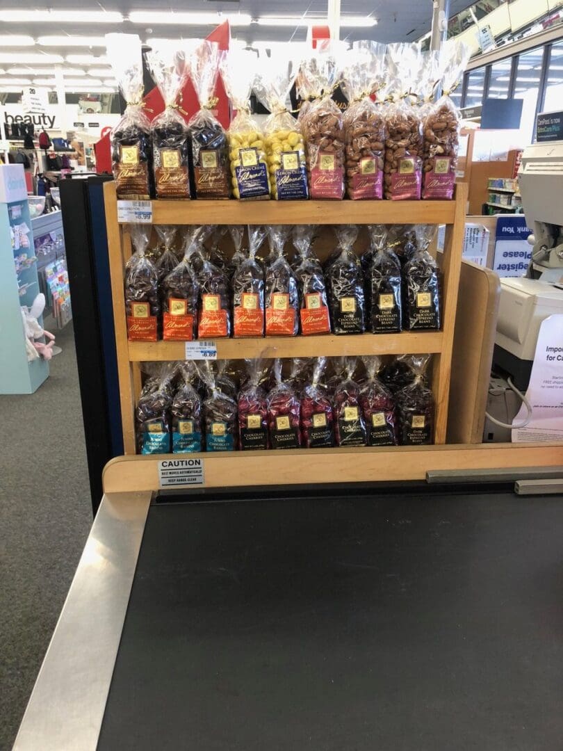 A display rack of various packaged breads near a checkout counter in a store, with a "caution" sign visible on the conveyor belt.