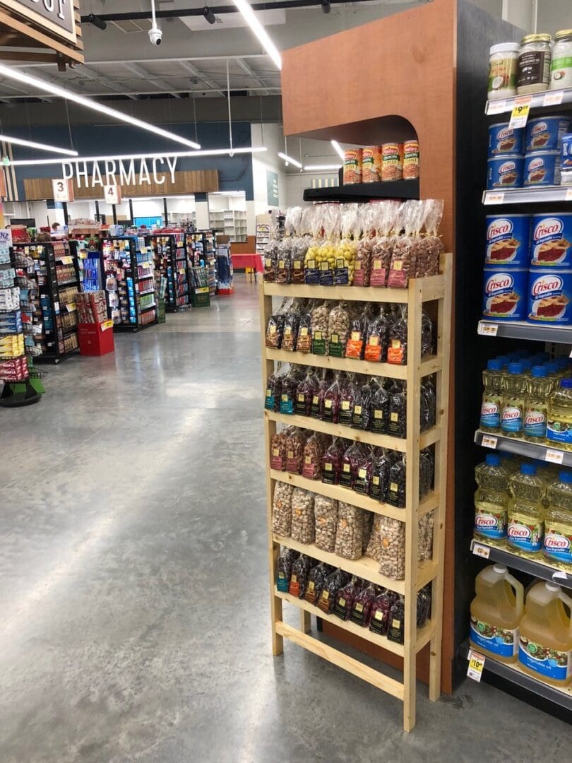 Aisle in a supermarket displaying a variety of food products including snacks and condiments, with a pharmacy sign in the background.