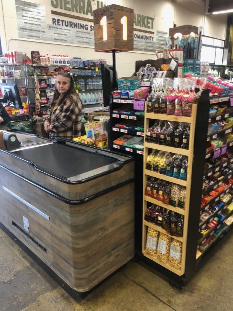 Woman standing at a grocery store checkout counter surrounded by various food items and snacks.