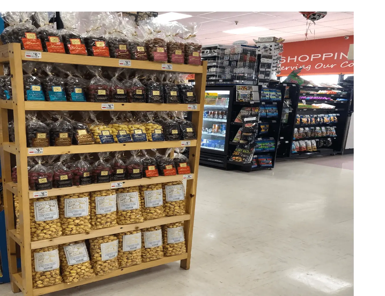 A grocery store aisle featuring a wooden display rack filled with various packaged nuts and dried fruits, with other snack shelves in the background.