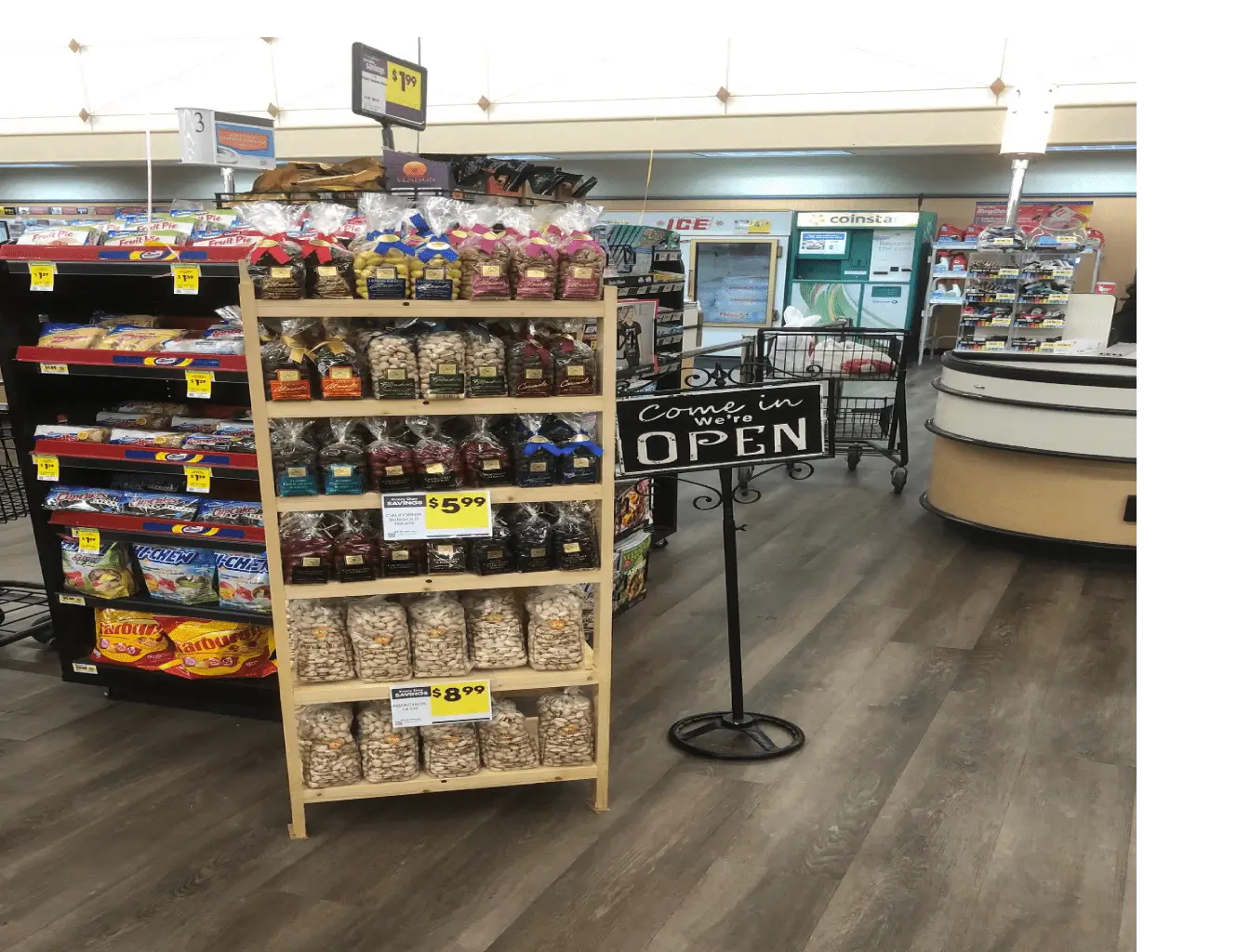 A retail store interior showing a display of assorted snack bags with prices, near an "come in we're open" sign, with shelves and a checkout counter in the background.